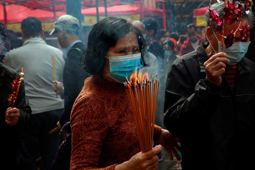 A middle-aged woman wears a mask and holds a bunch of incense sticks surrounded by other worshippers at a temple.