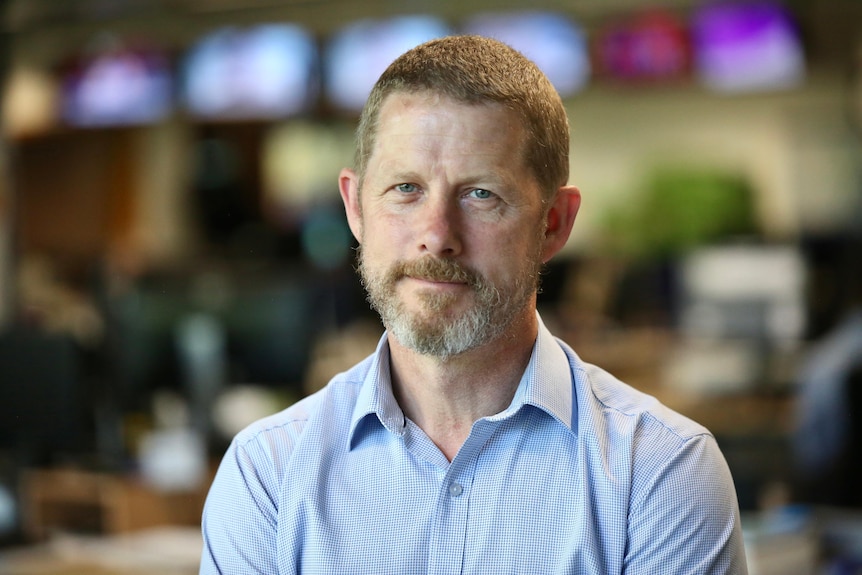 A lightly bearded man with short hair and a blue business shirt stands in a newsroom 