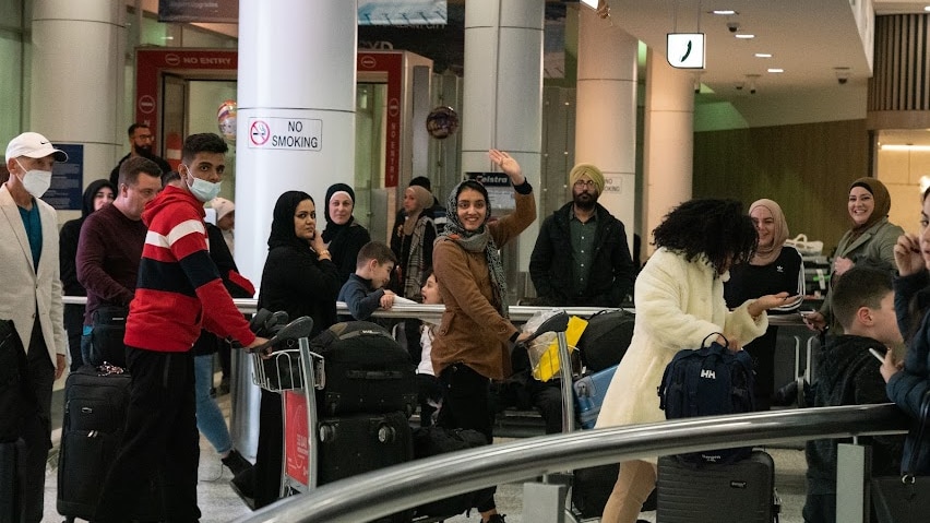 A young woman in a headscarf waves and smiles at the camera as she walks in an airport line