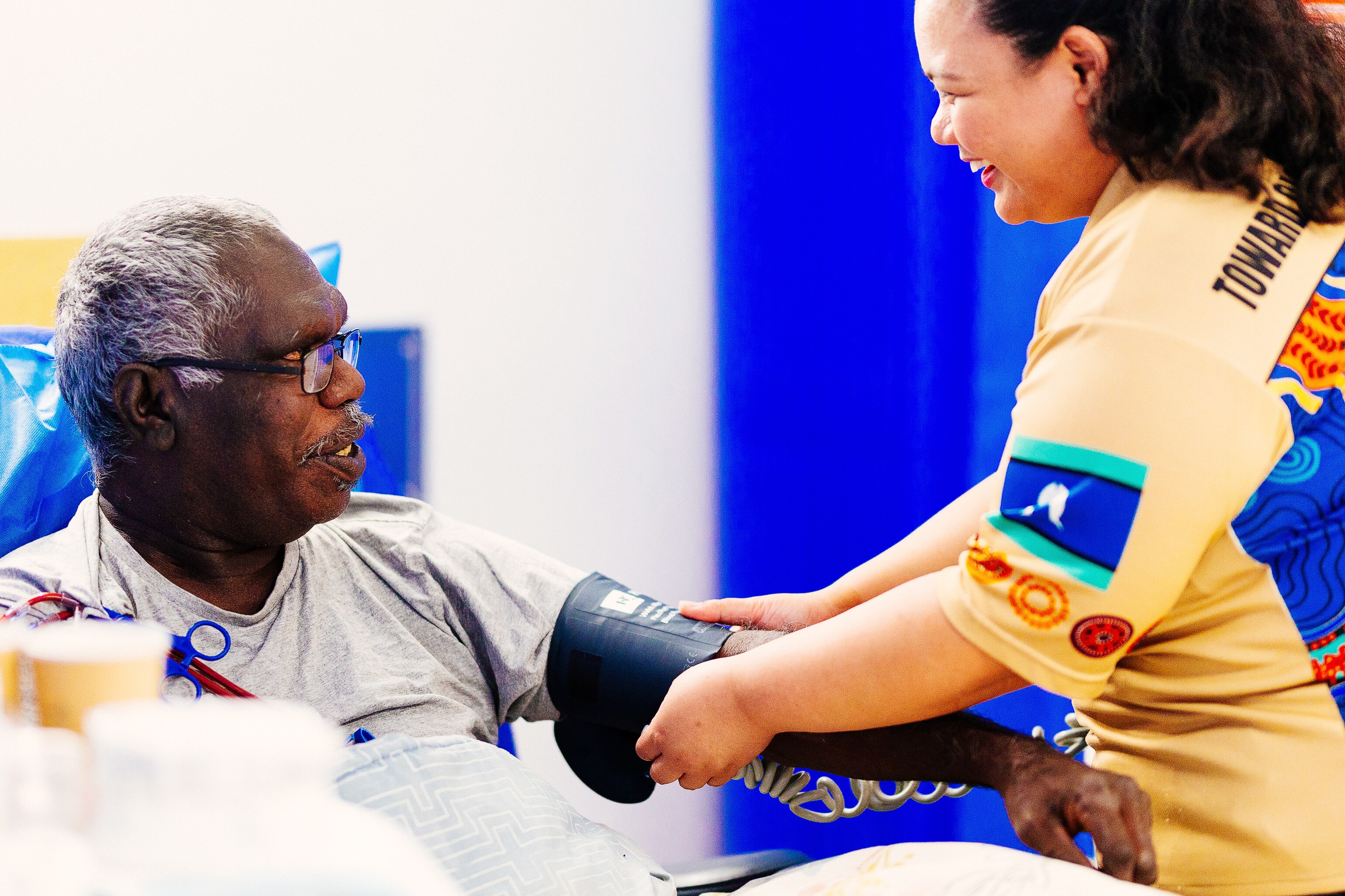 A woman wraps a blood pressure bandage around an Indigenous man's arm as he sits in a chair.