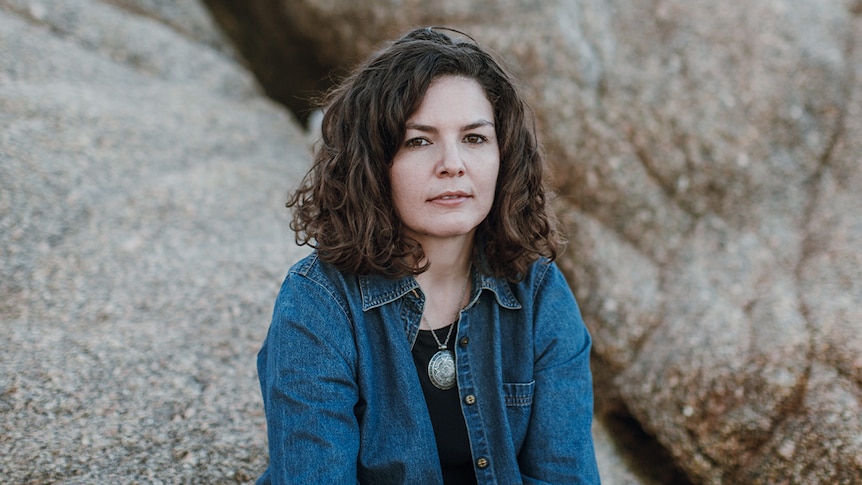 Eliza Hull sits outside and looks at the camera. She wears a denim jacket and large pendant necklace and shoulder length hair