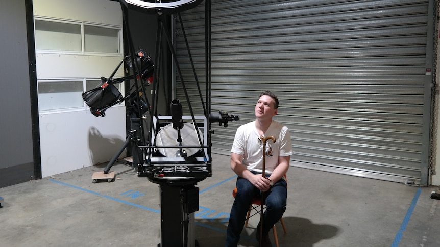 A young man sits on a chair and looks at a large telescope.