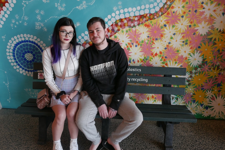 A young couple sit on a park bench in front of a colourful mural.