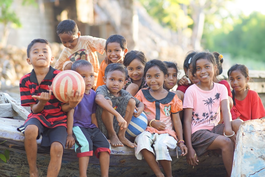 A group of children sit holding a ball and smiling near palm trees.