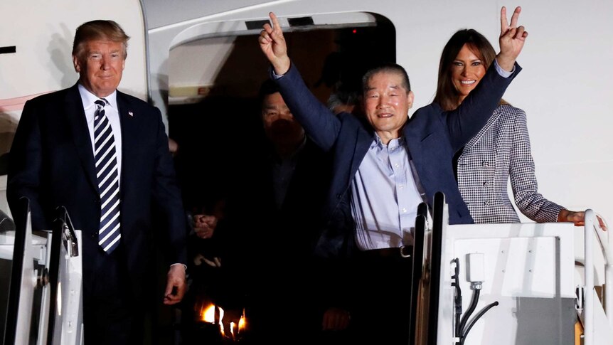 One of the Americans formerly held hostage in North Korea raises his arms and is accompanied by Donald Trump and Melania.
