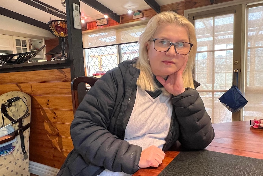 A serious blonde woman, wears glasses, puffer jacket, sits in a living room, leans upon a dining table, looks at the camera.