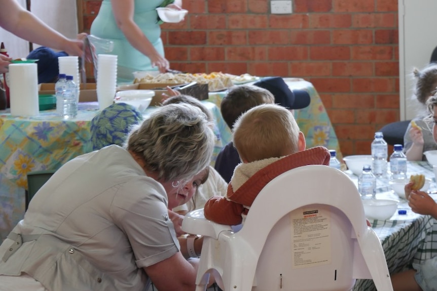 A woman kneels down to help young children with lunch
