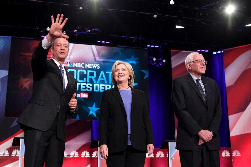 Martin O'Malley, Hillary Clinton and Bernie Sanders stand on the stage before the debate, with O'Malley waving to the audience.