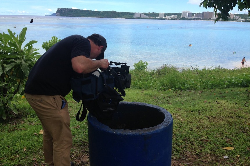 Greg Nelson resting camera on rubbish bin while filming a beach in Guam.