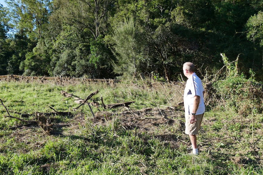 80 year old man stands looking at debris aftermath on his flood damaged property