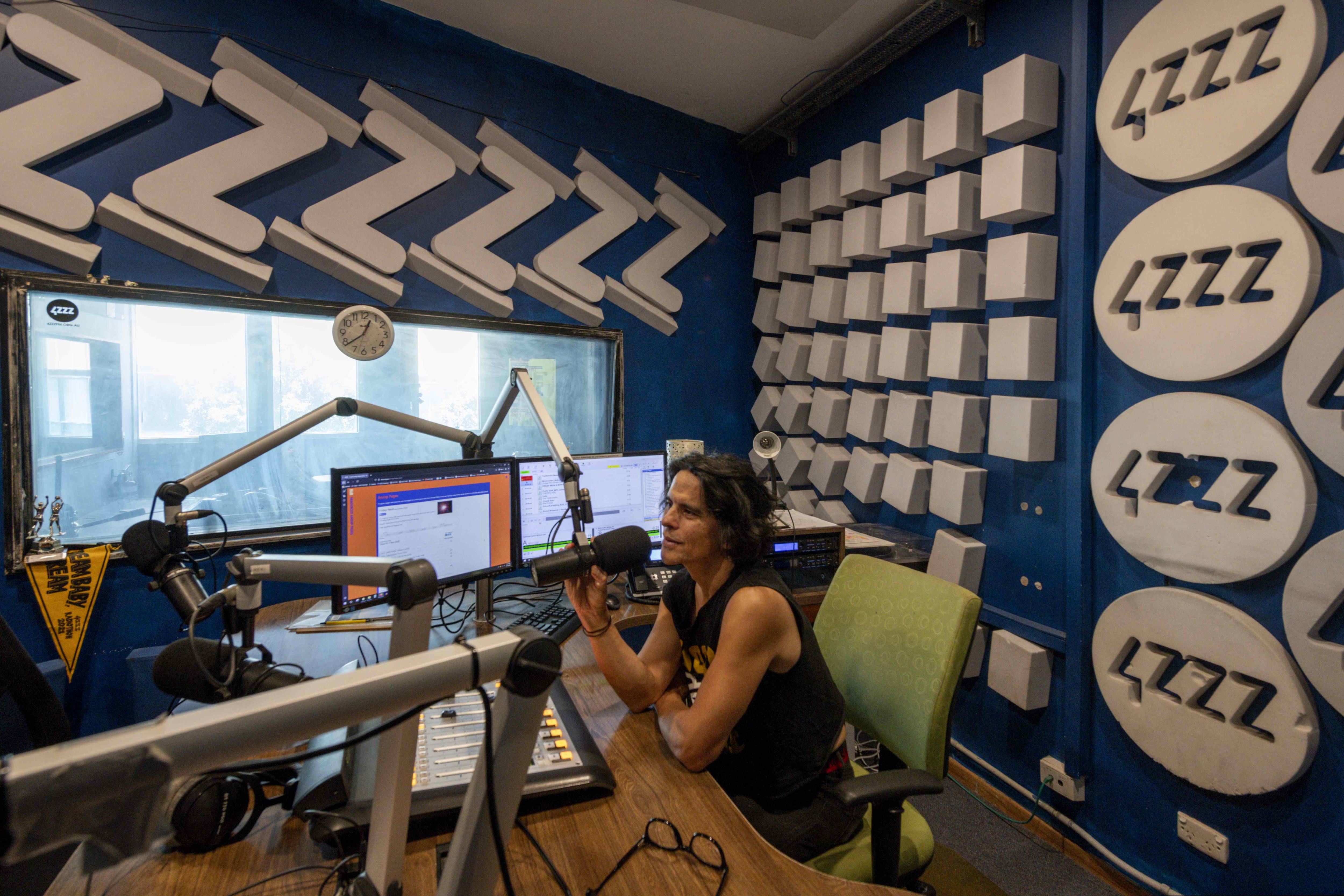 Long-haired man in sleevless top sits at radio desk with mic inside 4ZZZ community radio station