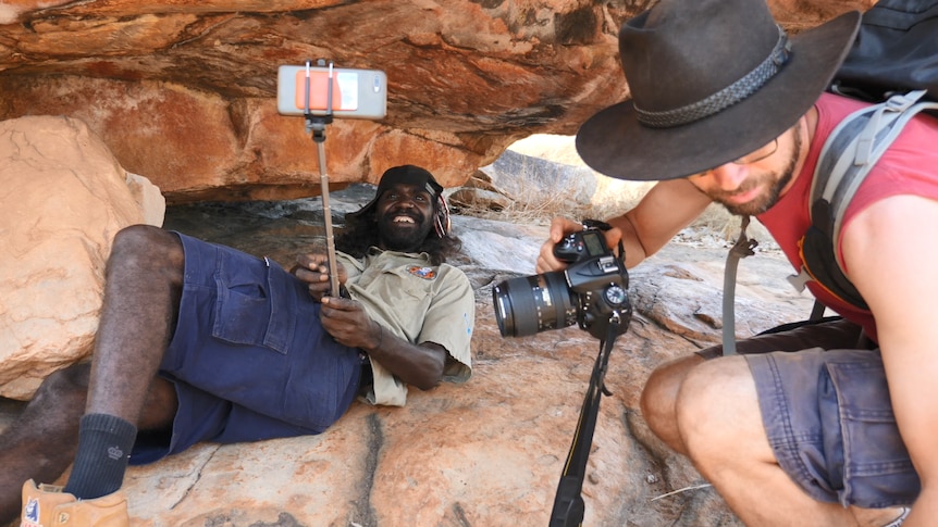 indigenous man sits under cave painting with selfie stick next to tourist