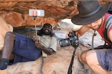 indigenous man sits under cave painting with selfie stick next to tourist