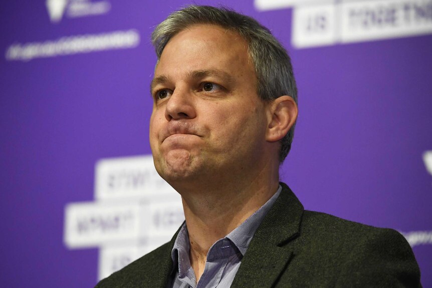 A headshot of Brett Sutton during a press conference against a purple background.