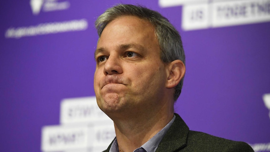 A headshot of Brett Sutton during a press conference against a purple background.