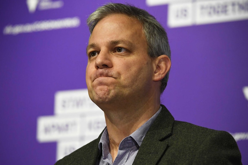 A headshot of Brett Sutton during a press conference against a purple background.