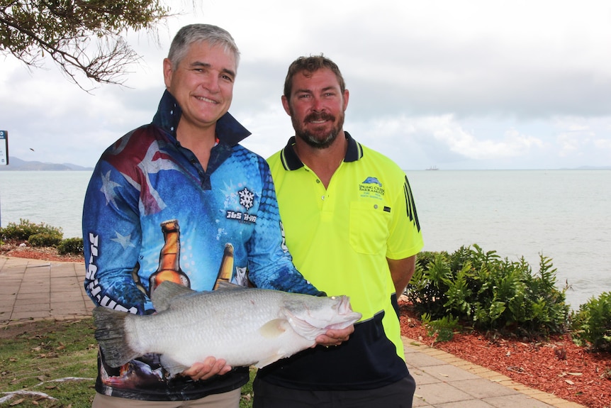 A man in a fishing shirt holds a barramundi smiling with a man in a fluro yellow shirt. Ocean in the background