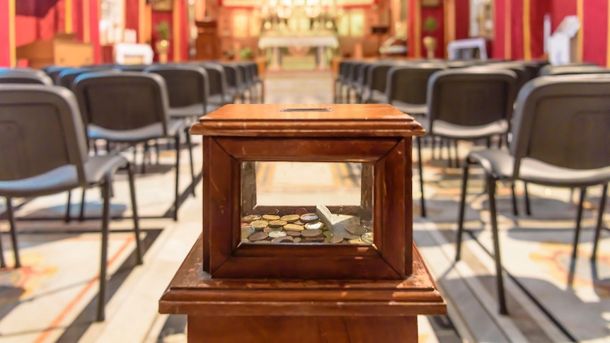A small wooden framed glass box with coins in it at centre. Behind, blurred, is large room with rows of chairs and Church altar.