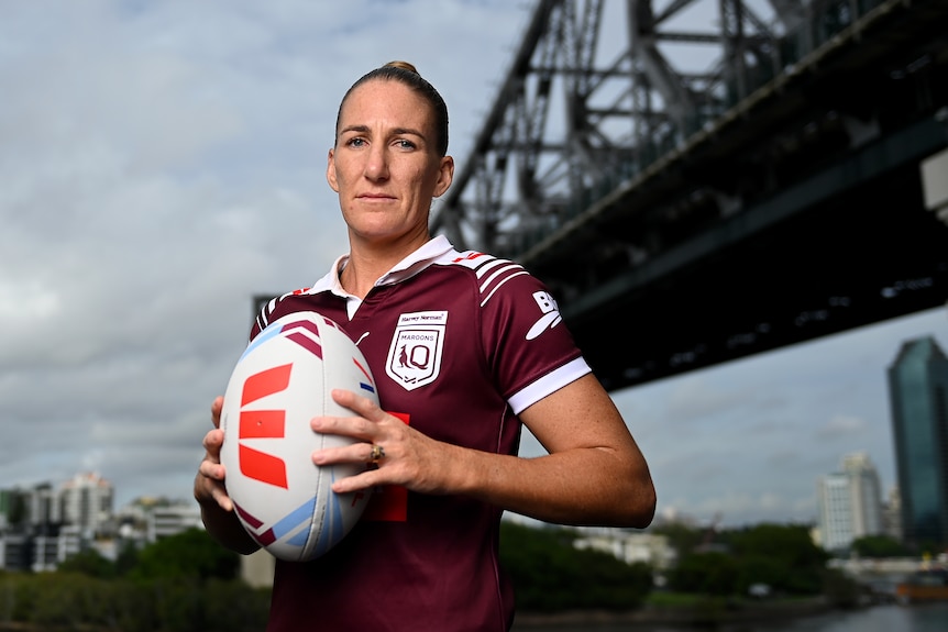 Female State of Origin player Ali Brigginshaw, wearing a Maroon jersey holding a football in front of a bridge