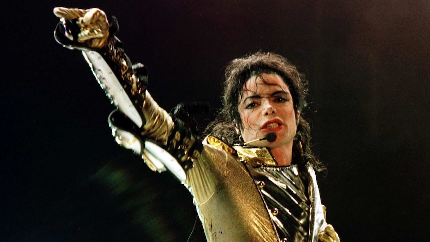 Michael Jackson, wearing a gold jacket and gloves, raises one arm up during a performance.