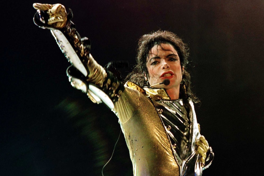 Michael Jackson, wearing a gold jacket and gloves, raises one arm up during a performance.