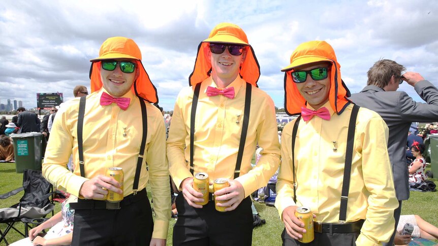 Three men wearing fluorescent orange legionnaires caps and pink bow ties at the races, drinking XXXX Gold stubbies.