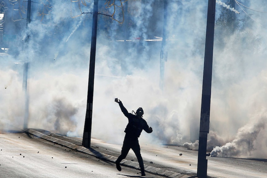 A Palestinian protester stands in the street and hurls stones into a fog of tear gas in Bethlehem.