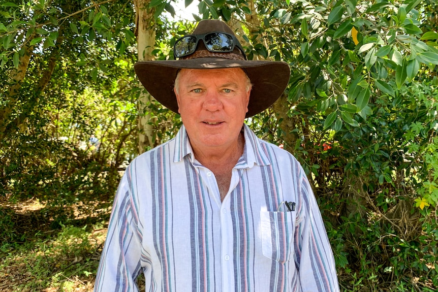 A male farmer wears a hat and looks at the camera as he stands next to a tree.
