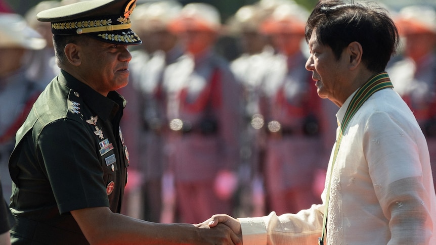 A man wearing a military uniform shakes hands with another man while a group of soldiers look on