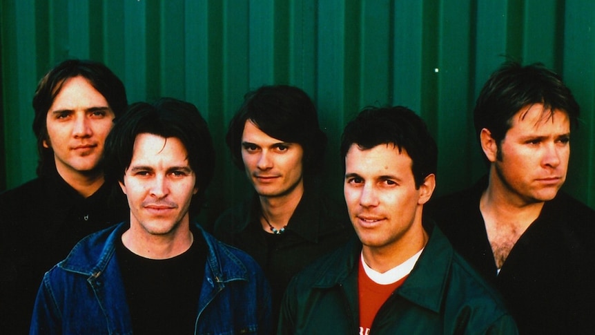 Five members of Brisbane rock band Powderfinger stand up against a green fence