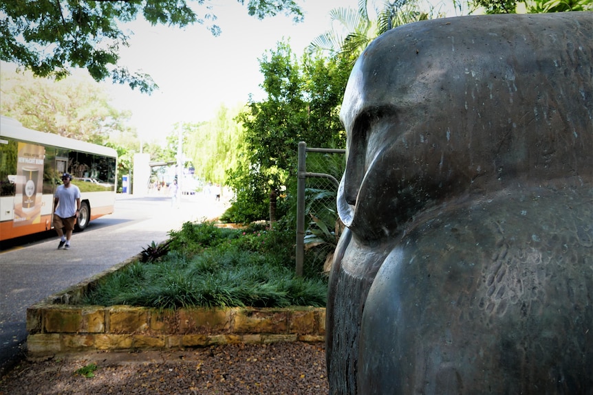 A close up of an owl sculpture's head with a bus and a man walking in the background.