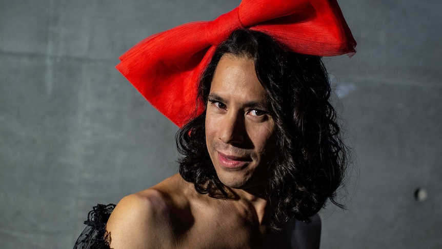 Man smiles to camera, wearing an oversized red bow on top of his head.