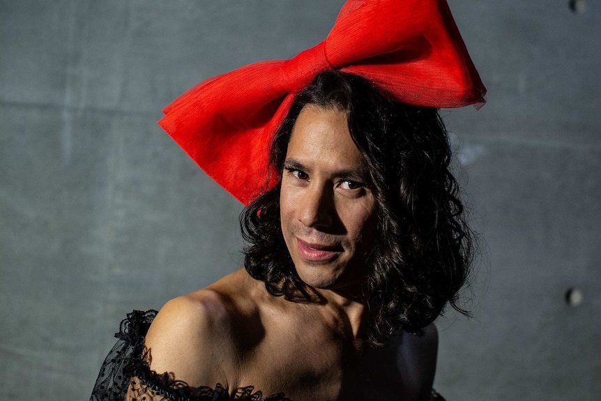 Man smiles to camera, wearing an oversized red bow on top of his head.