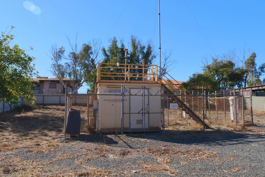 A small white structure surrounded by a security fence sits in the middle of a vacant allotment.