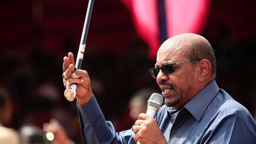 Sudan's President Omar al-Bashir speaks into a microphone while holding an ornate cane up in the air with one hand.