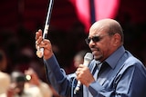 Sudan's President Omar al-Bashir speaks into a microphone while holding an ornate cane up in the air with one hand.