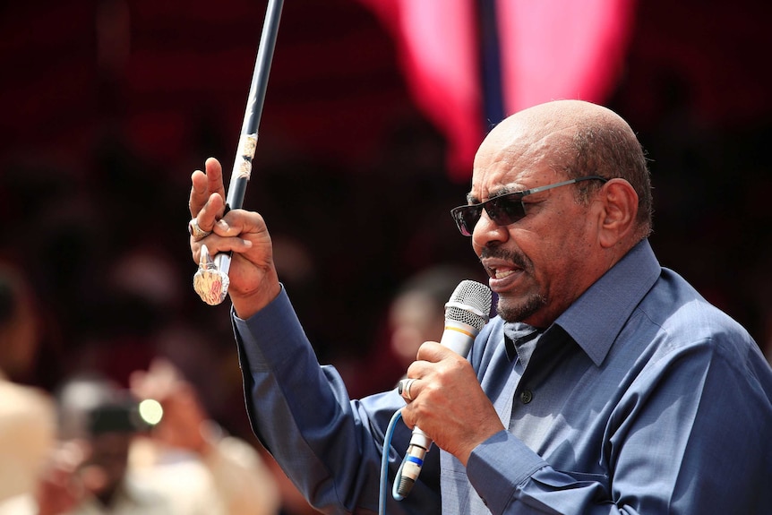 Sudan's President Omar al-Bashir speaks into a microphone while holding an ornate cane up in the air with one hand.