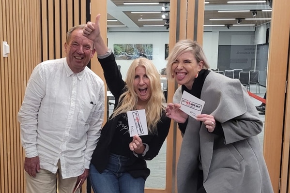 A smiling middle-aged man and two blonde women holding up campaign postcards 