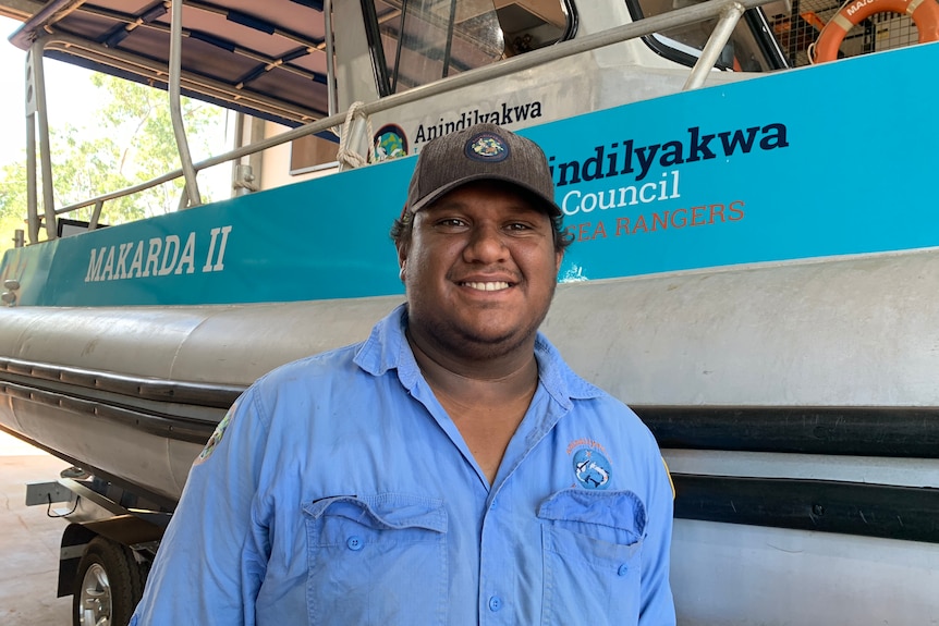 Anindilyakwa ranger Leyton Hastings in a blue polo shirt on land standing in front of a boat.