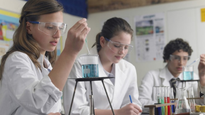 Three intent student scientists are dropping solution into a test tube over a burner