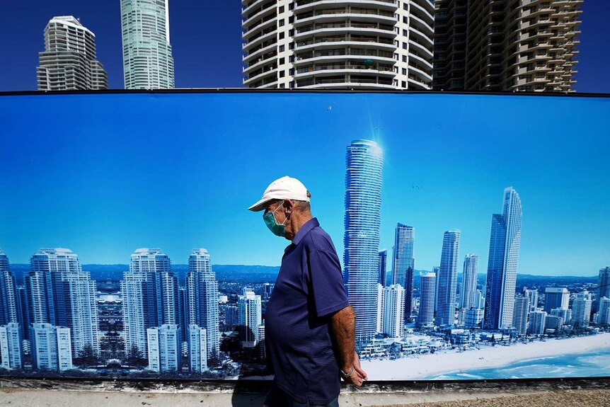 A man wearing a face mask walks past a sign with the Gold Coast skyline on it.