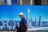 A man wearing a face mask walks past a sign with the Gold Coast skyline on it.