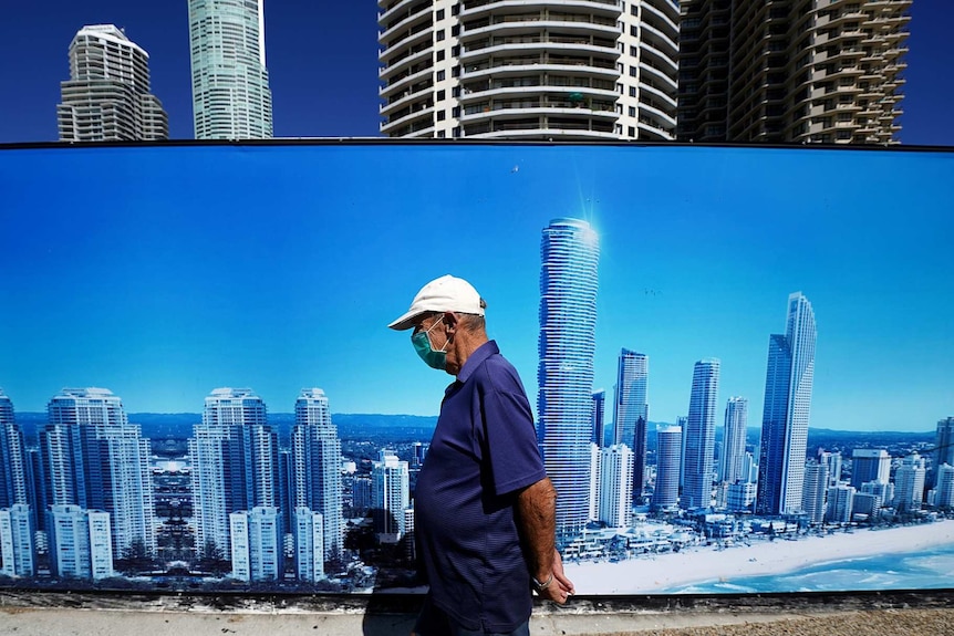 A man wearing a face mask walks past a sign with the Gold Coast skyline on it.