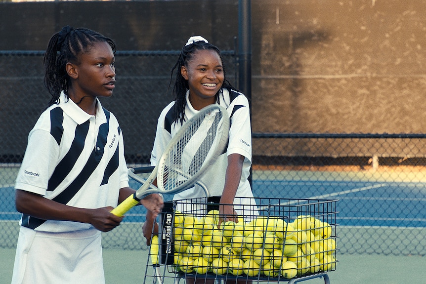 Two teen African American girls stand on a tennis court, one poised to return a hit, the other smiling near a basket of balls