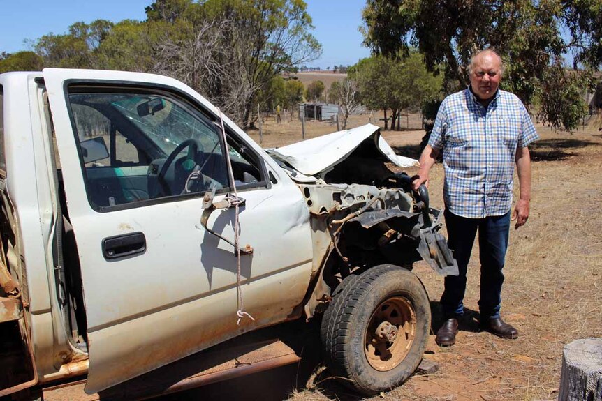A man in his 70's stands next to the wreckage of a white farm ute.