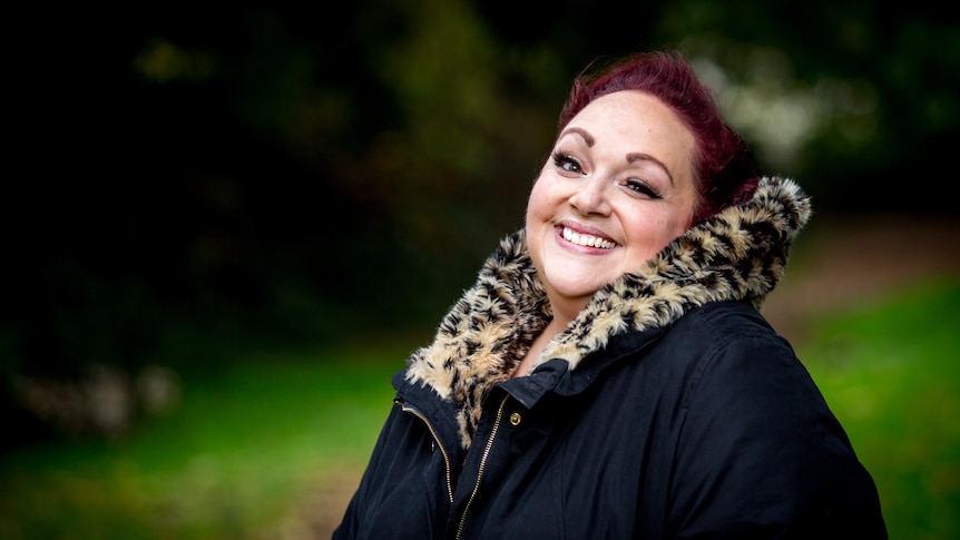 Helena Dix in a warm black coat with a fur neck-lining stands in a park and smiles at the camera.
