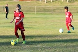 Two boys in red shirts kick a soccer ball across an oval.