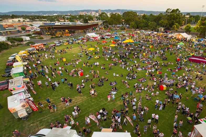 An oval with stalls around it and people in the middle with a community centre in the background