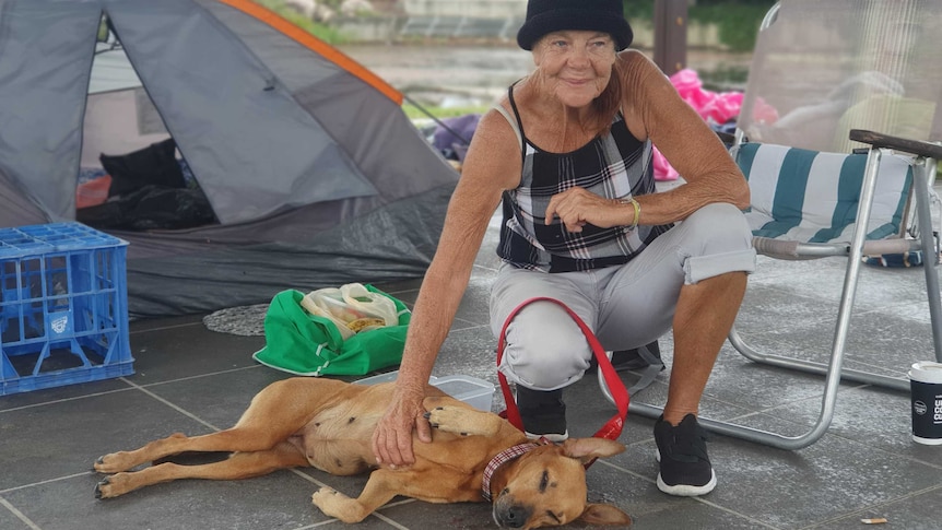 A homeless woman patting her dog with a tent behind her