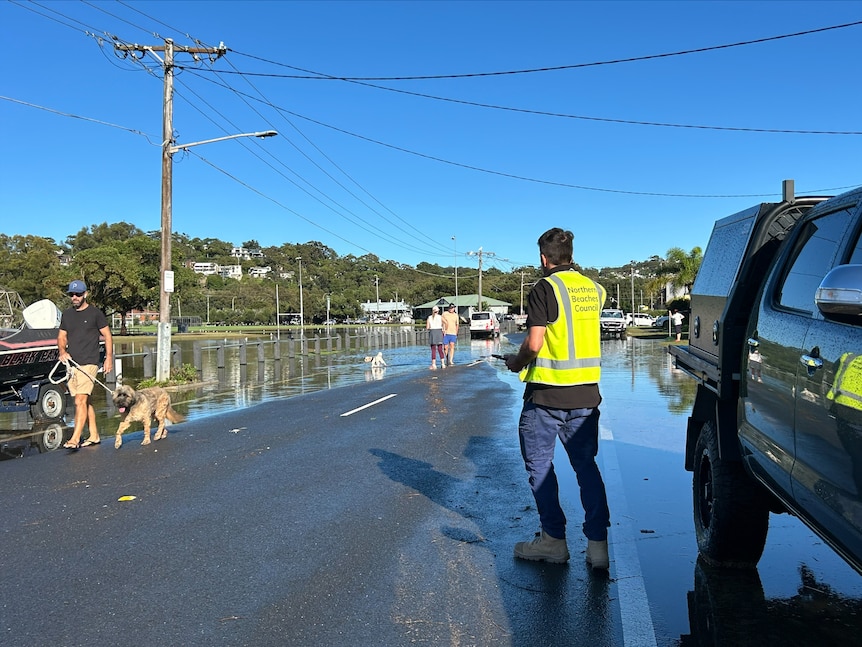 A flooded road with a rugby field in the background. A council worker and other people walking their dogs are in the foreground.